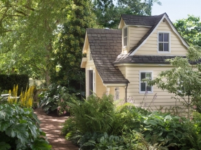 A charming playhouse cottage sits in a shaded perennial garden.