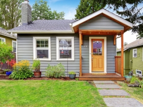 Small grey house with wooden deck. Front yard with flower bed and lawn